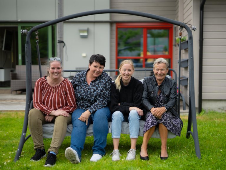 Fire kvinner sitter sammen i en hammock. De smiler og ser rett i kameraet. Et grått hus med en rød inngangsdør er synlig i bakgrunnen.